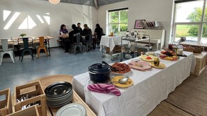 Um espaço amplo e interno com teto alto, mesas de madeira e uma longa mesa posta com comida.