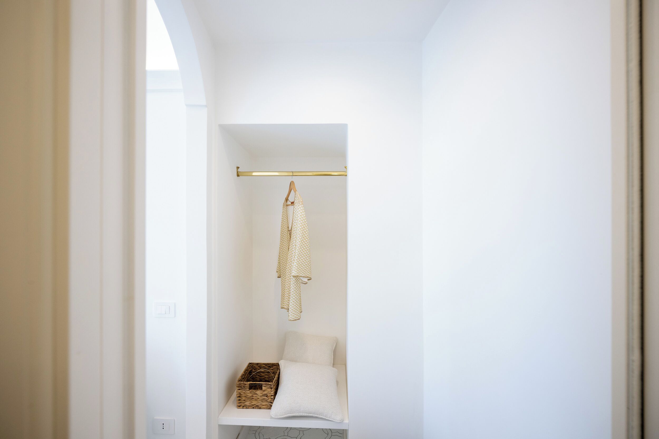 A narrow, white-walled bathroom with a hanging towel rack, a small bench with a basket, and a white pillow.