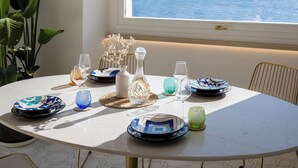 Mesa redonda de comedor con platos, vasos y un jarrón, frente a una pared blanca con un ventanal amplio que da al mar.