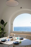 Mesa redonda de comedor con platos, vasos y un jarrón, frente a una pared blanca con un ventanal amplio que da al mar.