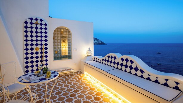 Una terraza con suelo estampado, una zona de estar con cojines, una mesa con un vaso y vistas al mar.