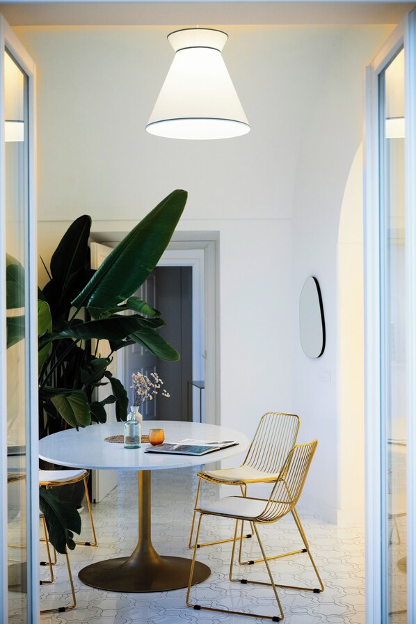 A round dining table with a white top and gold base, surrounded by two gold wire chairs. A large green plant is beside the table, and a white vase with flowers is on the table.