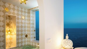 Una habitación con papel tapiz estampado, una lámpara en forma de estrella, una ventana con vista al mar y un objeto blanco decorativo sobre una mesa.