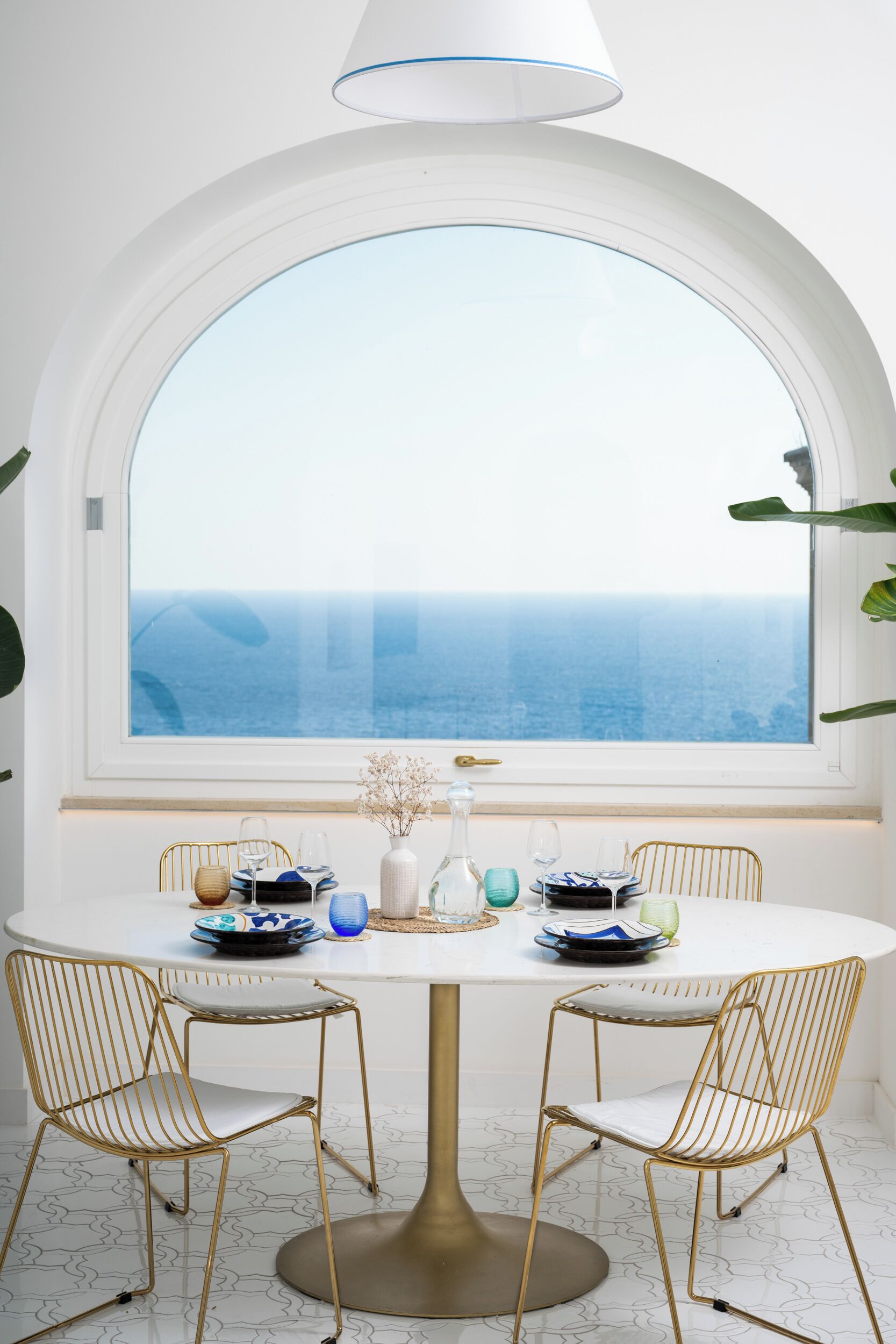 Una mesa de comedor con dos sillas de alambre dorado, un mantel blanco y vista al océano a través de un amplio ventanal.