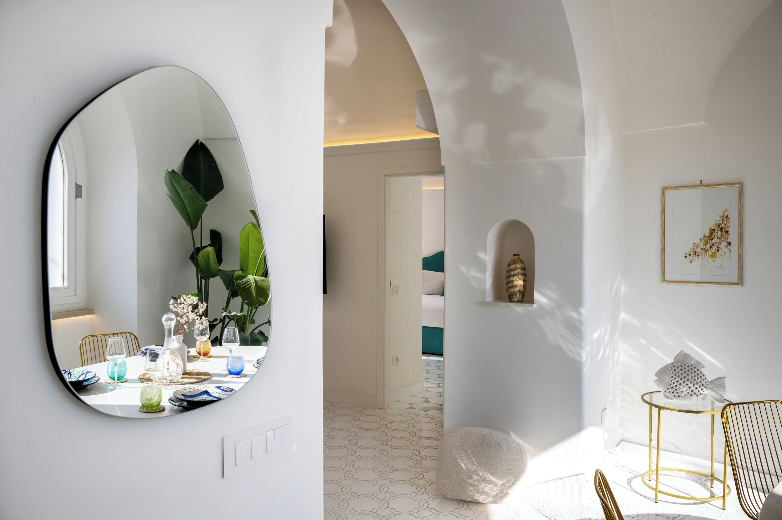 Ein modernes Interieur mit einem runden Spiegel, einem Esstisch mit Dekoration, einer weißen Wand mit einem gerahmten Kunstwerk und einer gemütlichen Sitzecke mit Stuhl und kleinem Tisch.