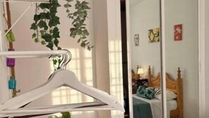 Une chambre avec un lit en bois, une plante suspendue, un cintre blanc et un sol carrelé.