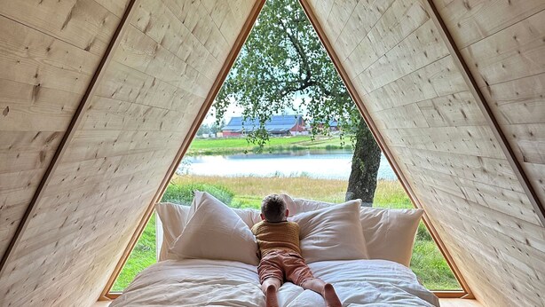 A child lying on a bed in a triangular wooden room with a view of a green landscape and a red building.