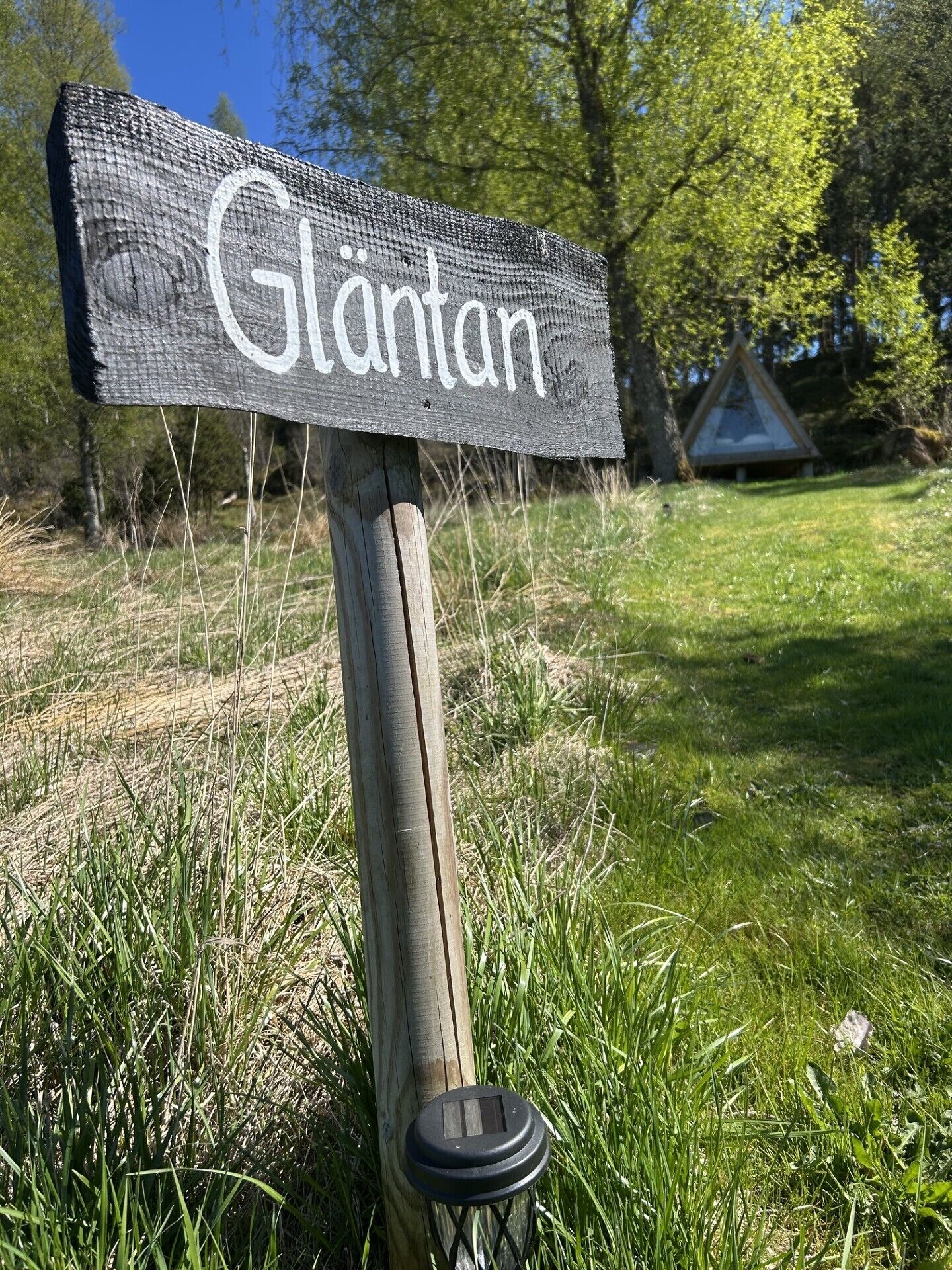 Puinen kyltti, jossa on valkoisilla kirjaimilla nimi »Glantan», metallinen tolppa ja ruohikkoalue, jonka taustalla näkyy puita.