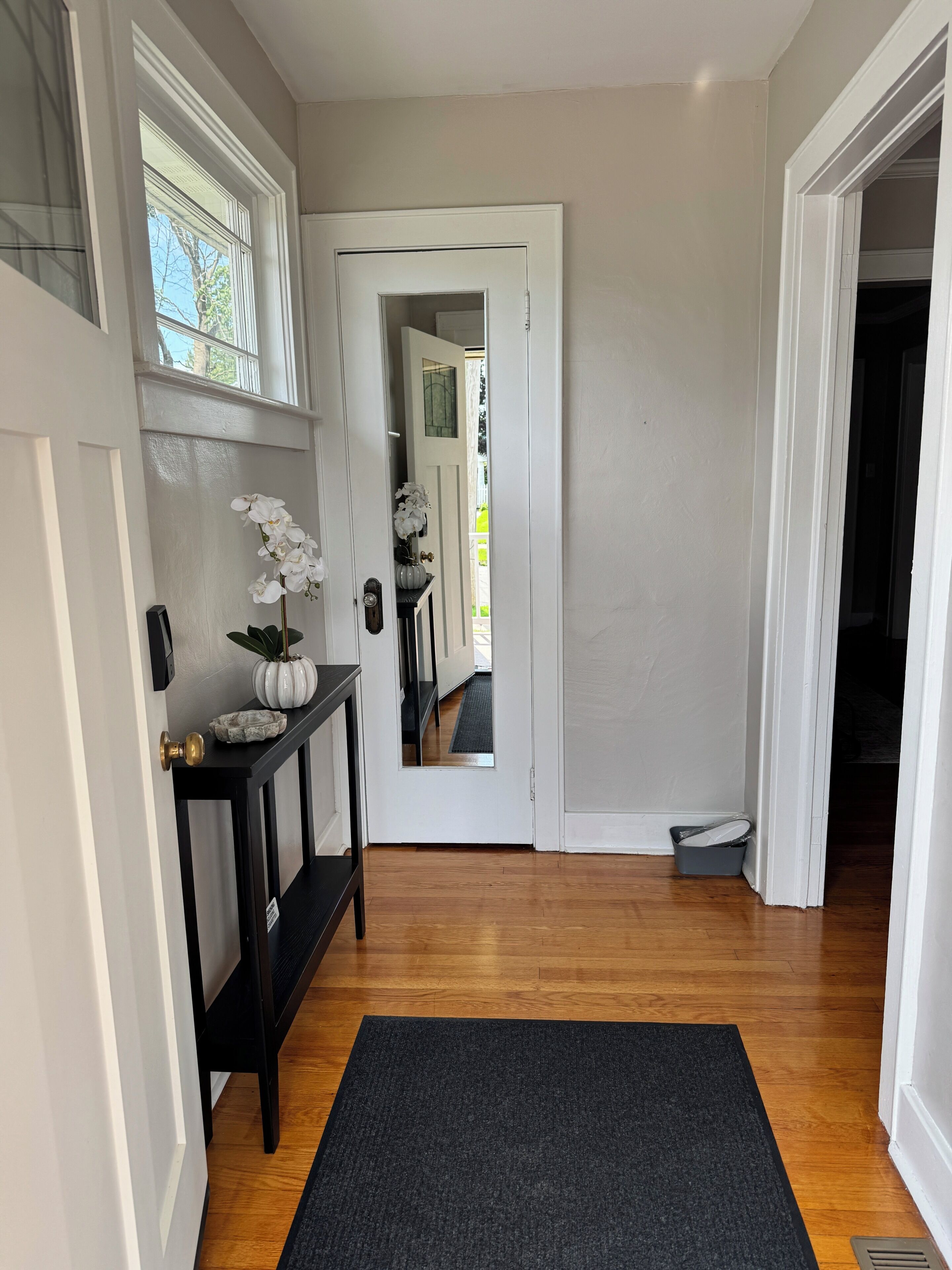 A hallway with a wooden floor, a black rug, a white door, and a small console table with a white vase and an orchid.