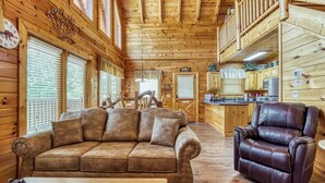 A cozy cabin interior with a wooden ceiling, a brown sofa, a leather recliner, and a dining area with wooden cabinets.