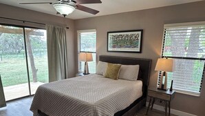 A bedroom with a large bed, two bedside tables with lamps, a ceiling fan, a window with blinds, and a framed picture on the wall. - Cool cottage on 30 acres between Blanco and Luckenbach (Blanco)