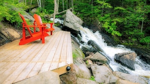 A wooden deck with two red Adirondack chairs overlooking a rocky stream in a forested area. - Loft Retreat on 24 acres with waterfall and hot tub (Highland Grove)