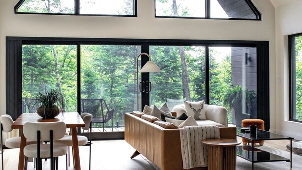 A spacious living area with a high ceiling, large windows offering a view of the outdoors, a wooden dining table with chairs, a sofa, and a coffee table. - Loft Retreat on 24 acres with waterfall and hot tub (Highland Grove)