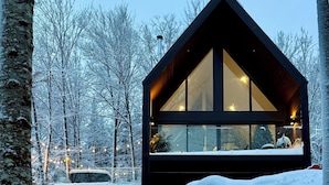 A modern cabin with large windows, a deck, and a snowy forest backdrop. - Loft Retreat on 24 acres with waterfall and hot tub (Highland Grove)