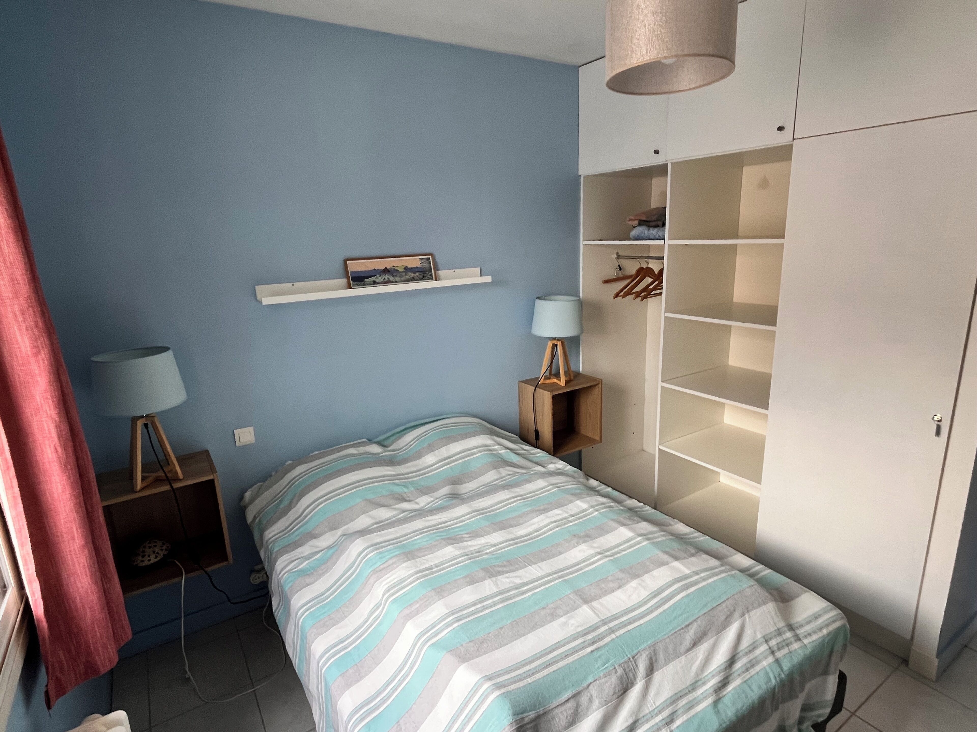 Un dormitorio con una cama, un armario, una estantería con un cuadro y dos lámparas de noche.