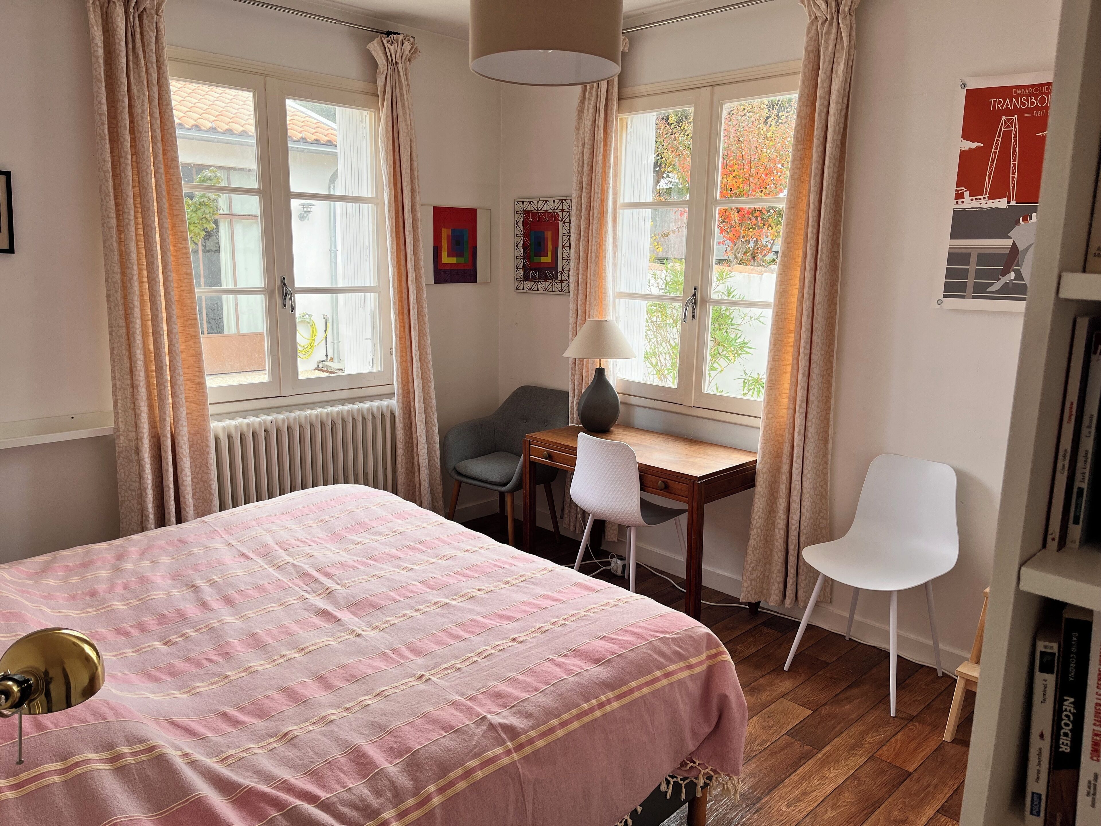 A bedroom with a bed, a desk, two chairs, a radiator, and a window with curtains.