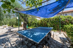 Une table de ping-pong installée dans un espace extérieur, protégée par une voile d’ombrage bleue, avec un sol en pierre et entourée de verdure.