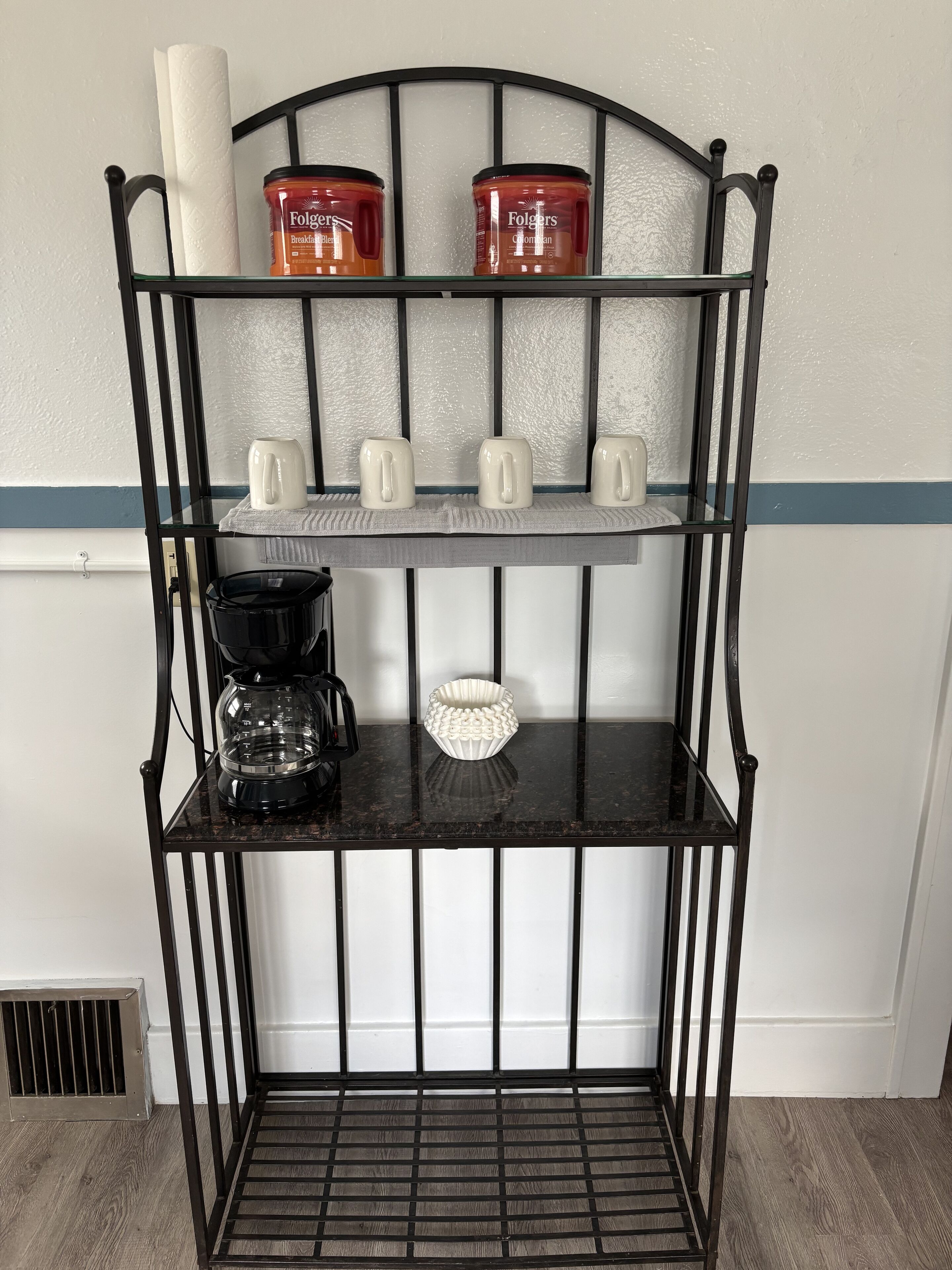 En svart metallhylla med tre våningar, där varje våning rymmer olika föremål, som till exempel burkar, muggar och en kaffebryggare.