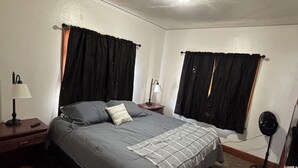 A bedroom with a bed, two nightstands, a lamp, a fan, and a window with curtains. - Charming 1-bedroom Duplex with AC, WiFi, Gym access etc... (Rawlins)