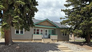 A single-story building with a green roof and beige siding, featuring a front door and windows, surrounded by gravel and trees. - Charming 1-bedroom Duplex with AC, WiFi, Gym access etc... (Rawlins)
