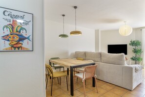 A living room with a sofa, a dining table with chairs, pendant lights, a flat-screen TV, and a framed picture on the wall.