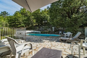 Zona de piscina al aire libre con sillones de descanso y una mesa, rodeada de suelo de piedra y árboles.