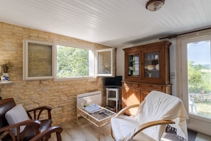 A room with stone walls, wooden furniture, a television, and a view of greenery outside.
