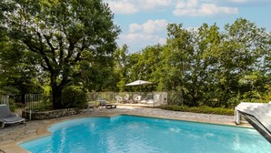 Une piscine entourée d’arbres et dotée d’une terrasse en pierre, avec des chaises longues et une table.