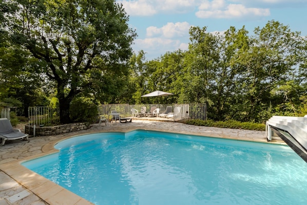 Una piscina con sillas de descanso y una mesa, rodeada de árboles y un patio de piedra.