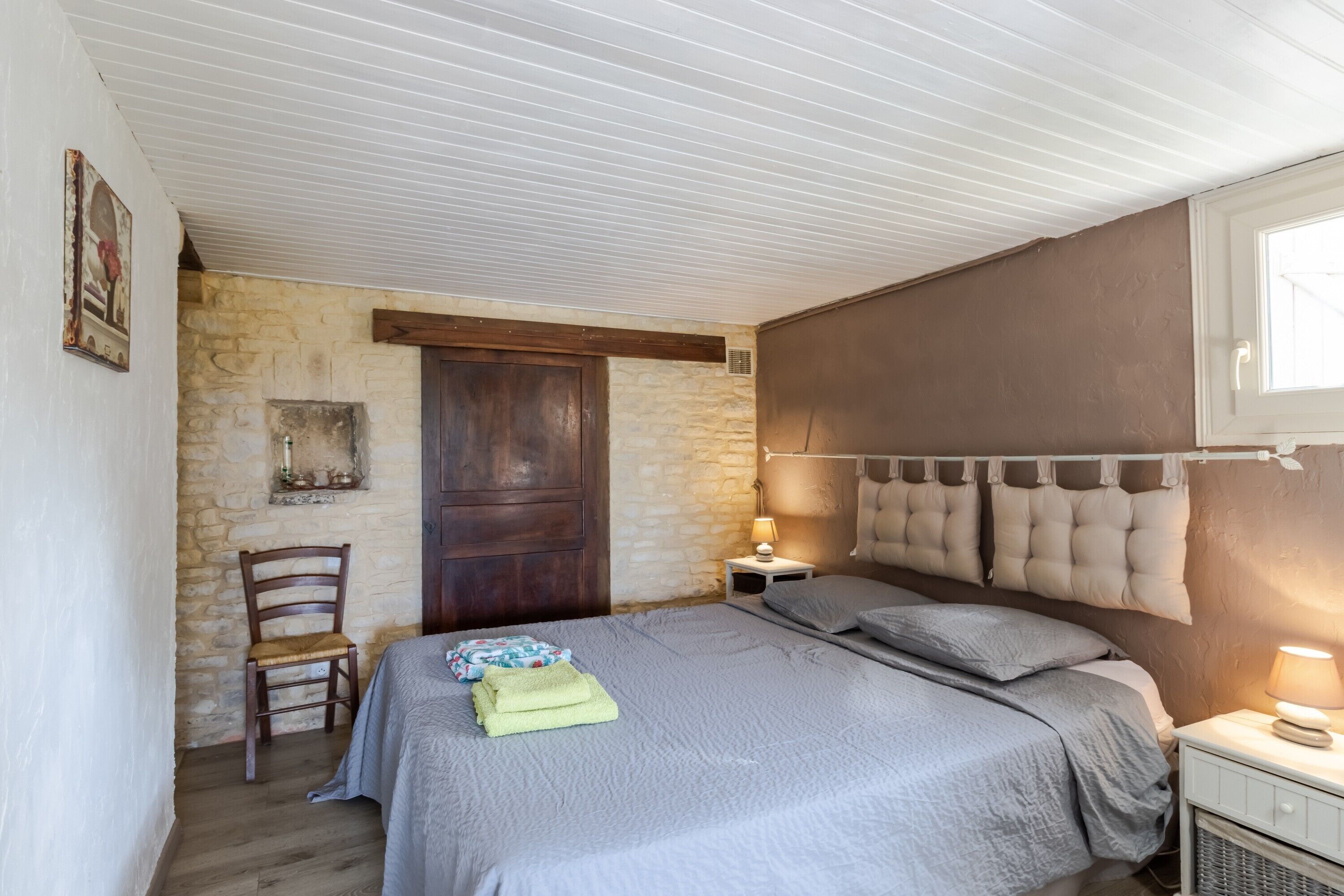 A bedroom with a bed, a chair, a nightstand, a lamp, a wooden door, and a stone wall.