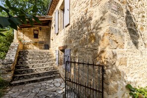 Un bâtiment en pierre avec un escalier menant à une porte, un portail en métal et un mur en pierre orné d’une plaque numérotée.
