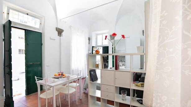 A room with a dining table, chairs, a green door, a white wall, a window, and a bookshelf with various items.