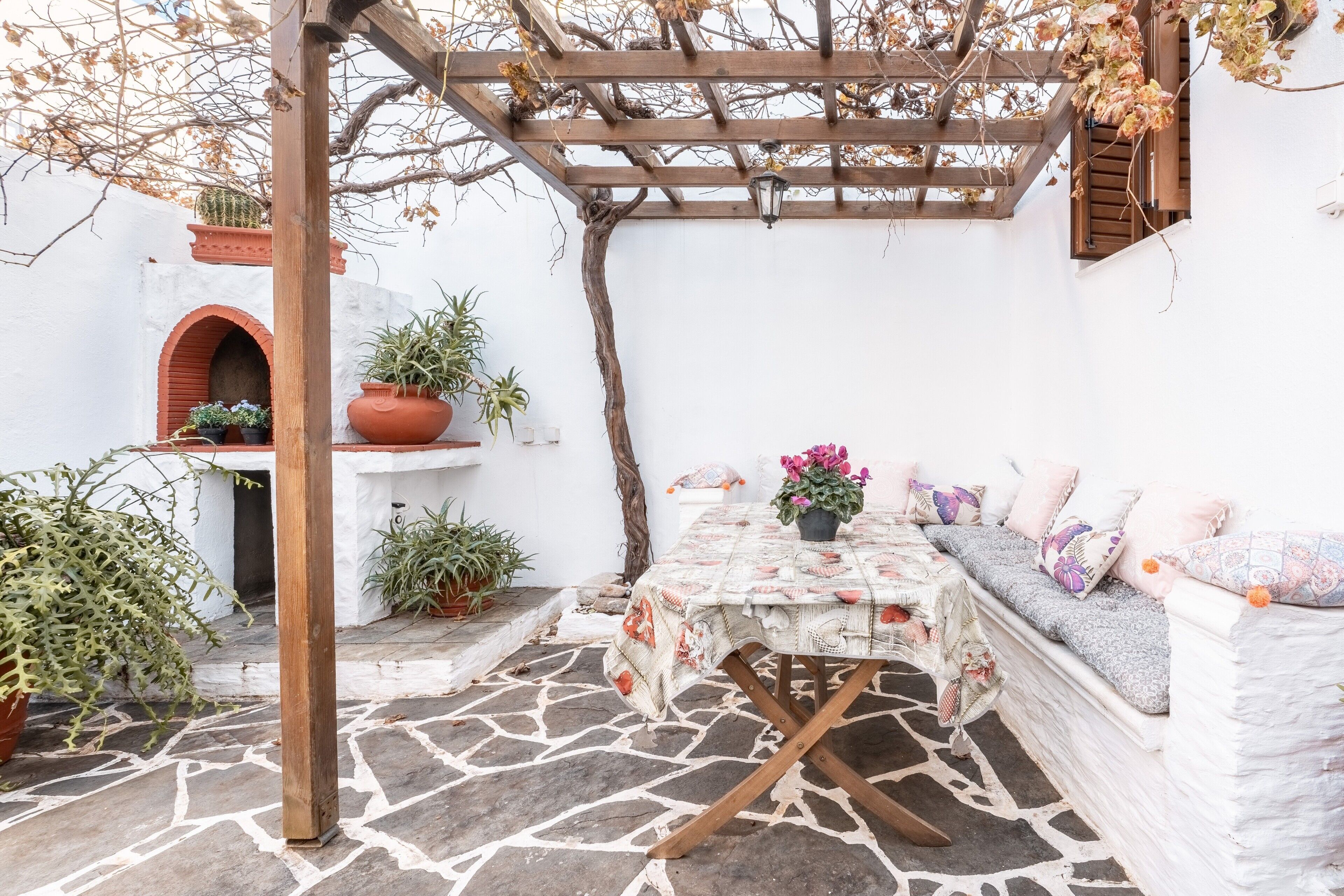 Un patio cubierto con una mesa y asientos, rodeado de plantas en macetas y una pared blanca.