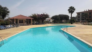 Una piscina rettangolare con acqua cristallina e azzurra, circondata da un solarium in cemento. Si vedono lettini e un piccolo edificio con tetto rosso sullo sfondo.