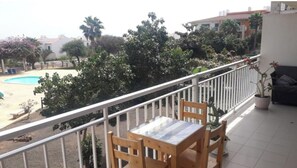 Ein Balkon mit Holztisch und -stühlen, einem Blumentopf und Blick auf ein Schwimmbecken sowie die umliegende Bepflanzung.