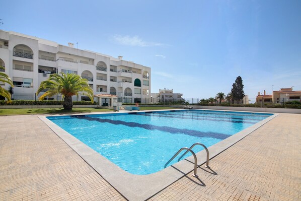 Una piscina con agua cristalina azul, rodeada por una terraza con baldosas, y un edificio de varios pisos con balcones al fondo.
