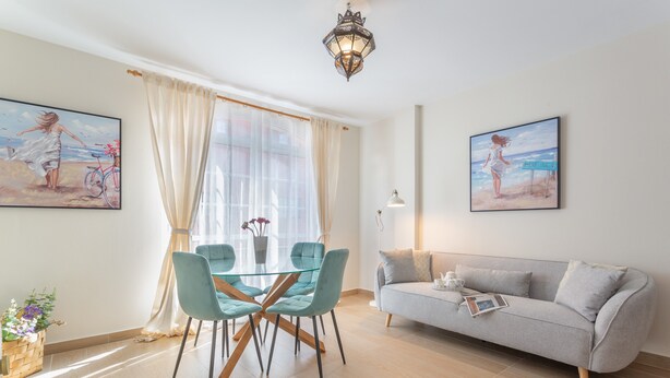 Een moderne woonkamer met een glazen eettafel, twee stoelen, een grijze bank en twee aan de muur hangende schilderijen.
