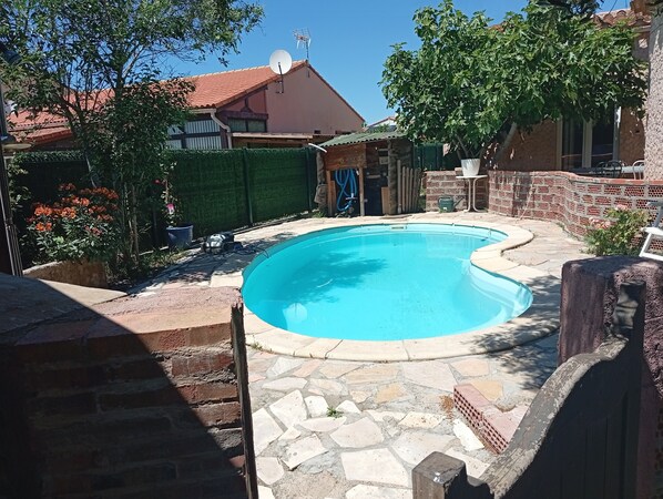 Piscina com deck de pedra, rodeada por um muro de tijolos e vegetação.