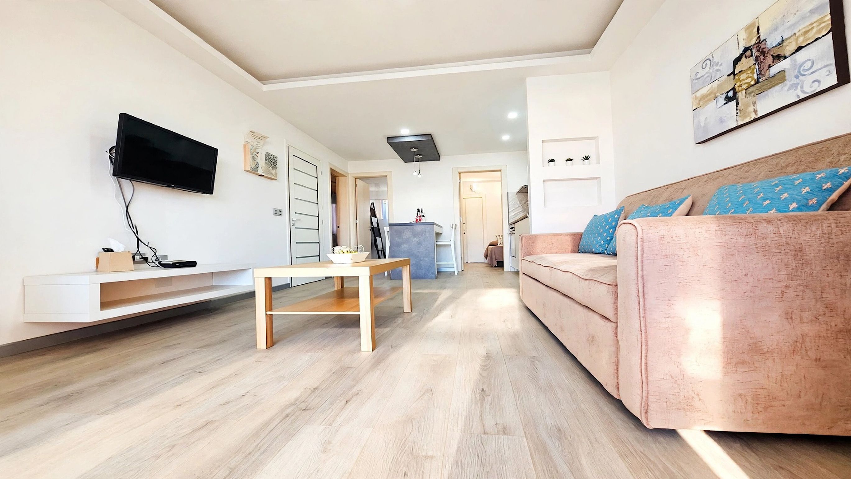 A modern living room with a flat-screen TV, a wooden coffee table, a sofa with patterned cushions, and a dining area with a small table and chairs.