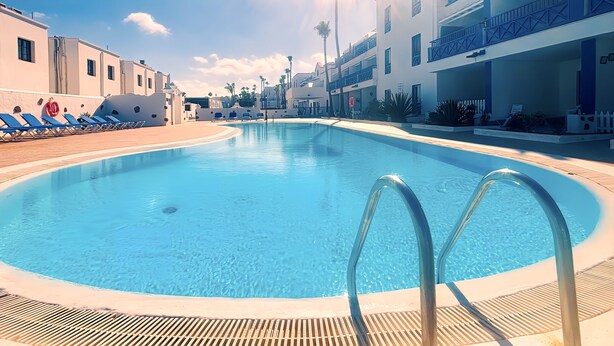 A swimming pool with sunlit water, lounge chairs, and modern buildings in the background. - Apartment Bellissima With Pool and Wi-fi, 200m From the sea in Puerto del Carmen (Tías)