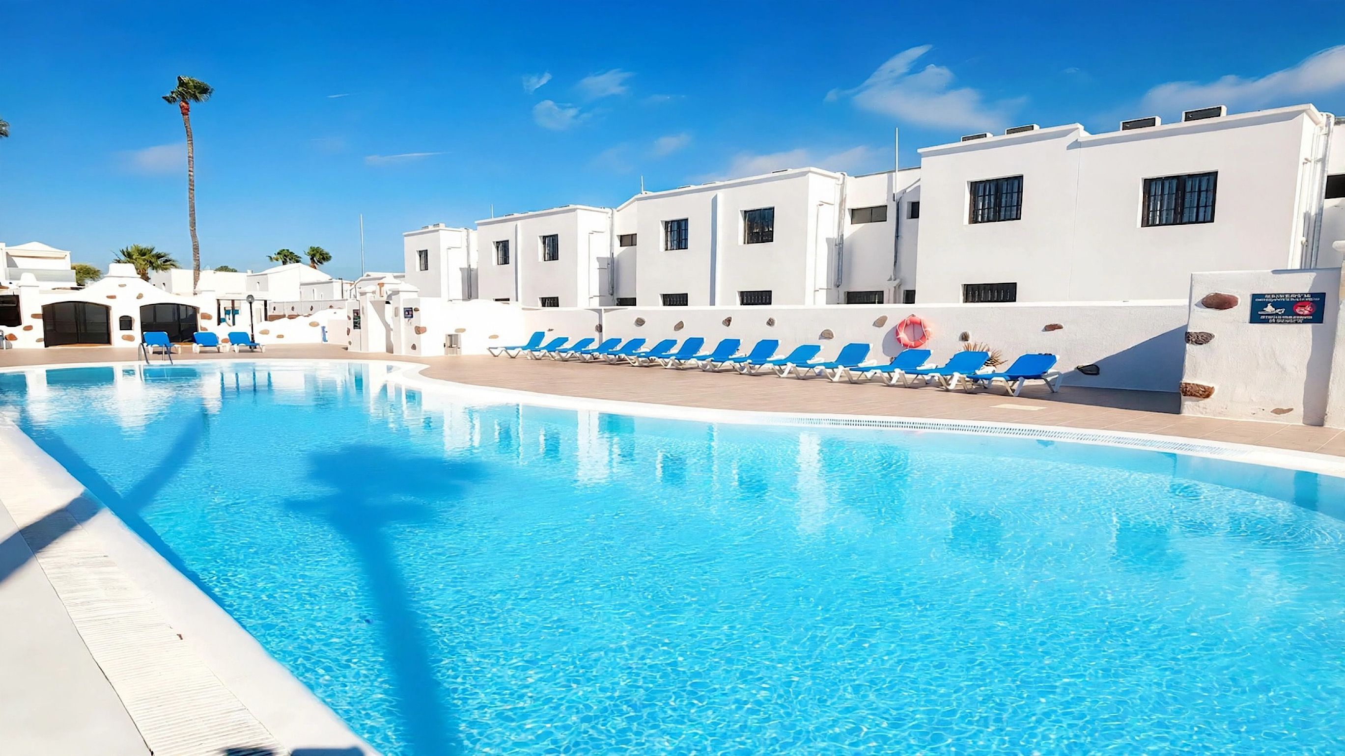 Une piscine à l’eau bleue, entourée de bâtiments blancs dotés de fenêtres, avec un palmier en arrière-plan.