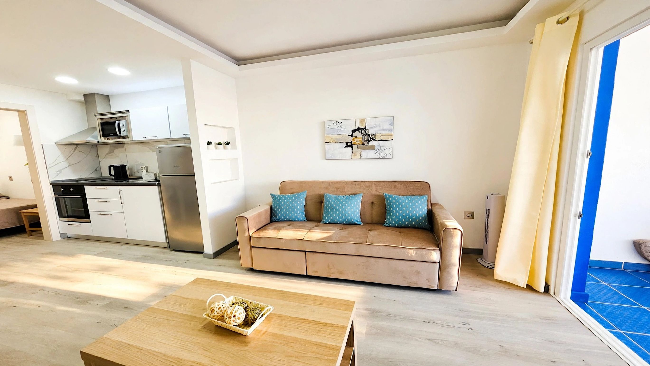Een knus woongedeelte met een kitchenette, een bank met gestreepte kussens, een houten salontafel en een vloer van blauwe tegels.