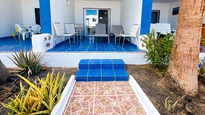 A covered outdoor seating area with white chairs and a blue-tiled floor, leading to a building entrance. - Apartment Bellissima With Pool and Wi-fi, 200m From the sea in Puerto del Carmen (Tías)