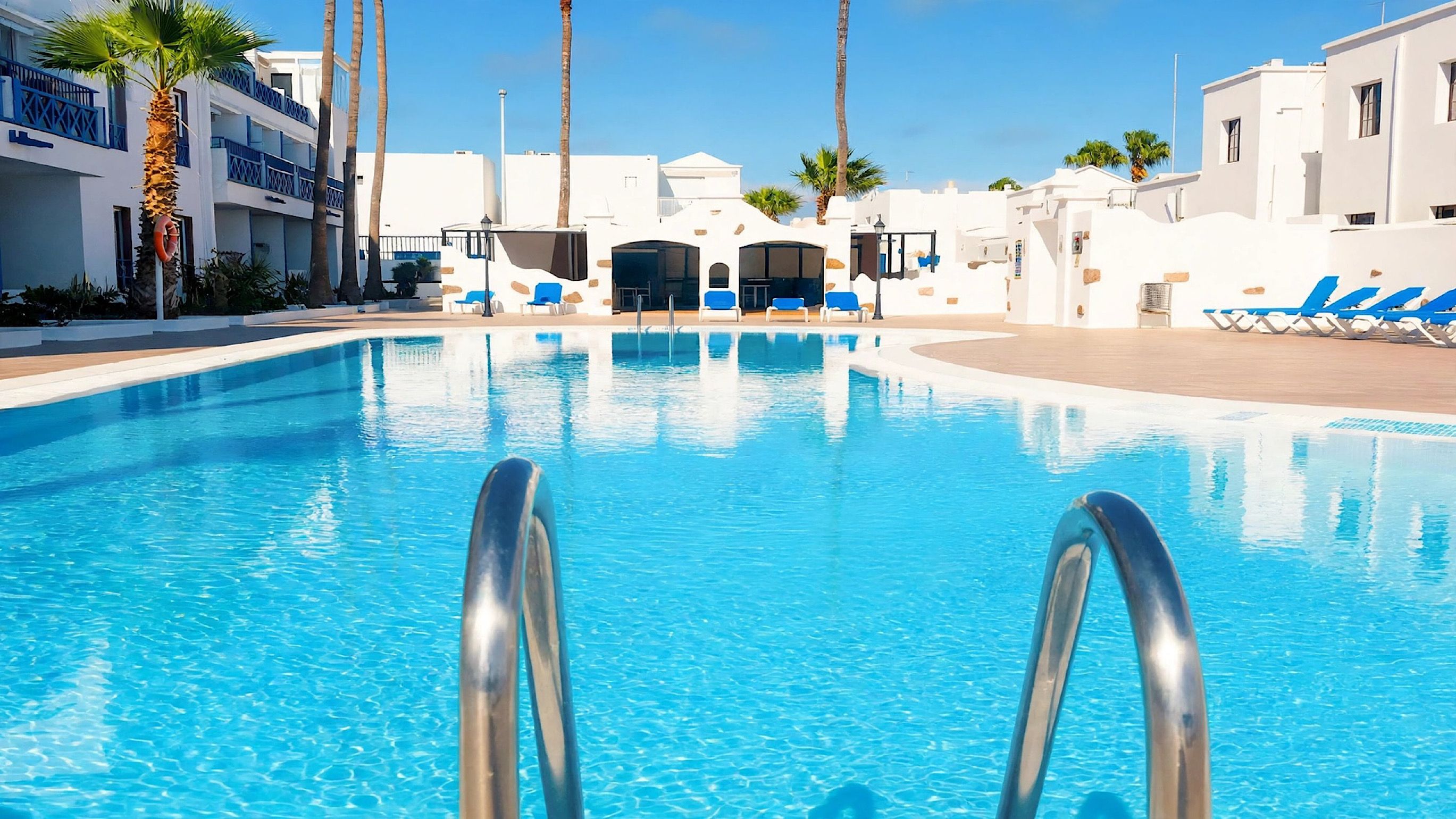 A swimming pool with clear blue water, surrounded by white buildings with blue accents, and a few palm trees.