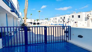 A rooftop pool area with a blue fence, white buildings, and palm trees in the background. - Apartment Bellissima With Pool and Wi-fi, 200m From the sea in Puerto del Carmen (Tías)