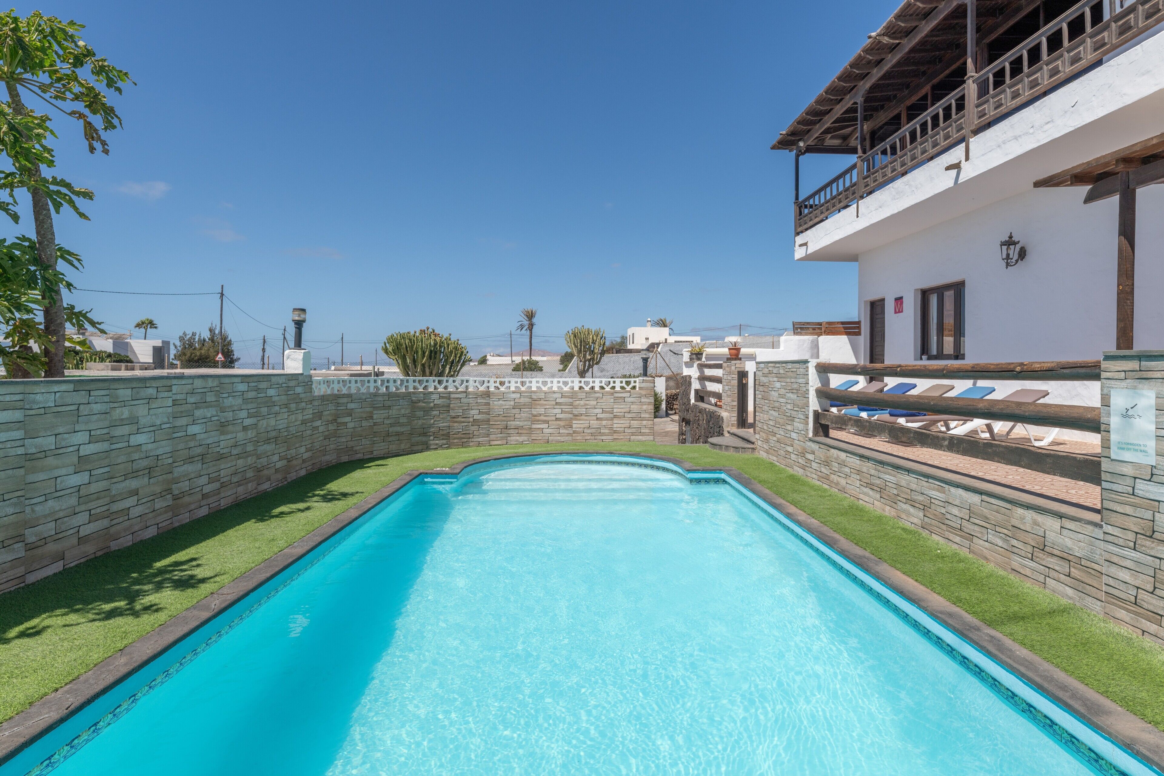 Ein Swimmingpool mit Steinmauer und Rasen, ein Balkon mit Liegestühlen und ein klarer, blauer Himmel.
