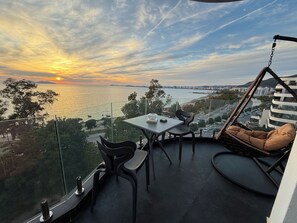 A balcony with a table and chairs, a hanging swing, and a view of the sunset over the water.