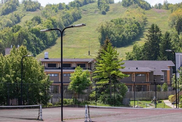 A tennis court with a net, surrounded by a fence, and a building with multiple windows in the background. - Slopeside Sanctuary (Blue Mountain)