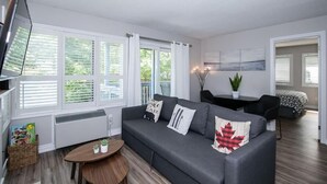 A modern living room with a grey sofa, a coffee table, and a TV. There are large windows with blinds, a small table with plants, and a painting on the wall. - Slopeside Sanctuary (Blue Mountain)