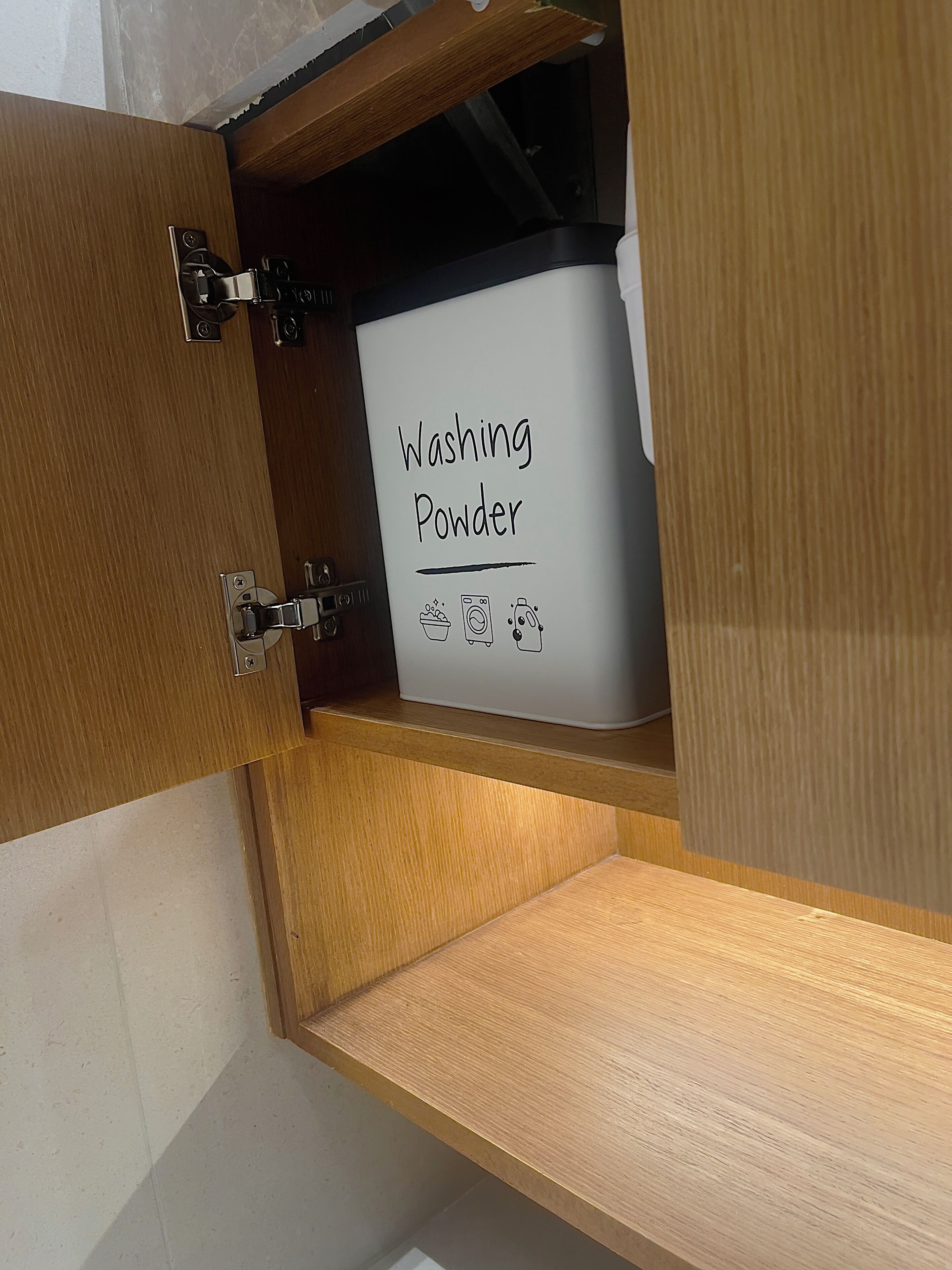 A wooden cabinet with a drawer containing a white box labeled 'Washing Powder' and a small image of a washing machine.